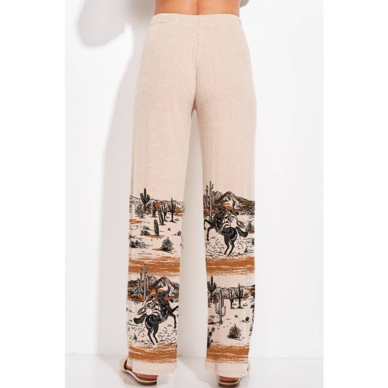 A pair of wide-leg knit pants in a light beige color, featuring a western desert print with cacti, mountains, and horse riders near the lower legs, shown on a model from the back.
