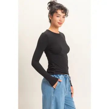 A woman modeling a fitted black long-sleeve crewneck top paired with light blue jeans, standing against a plain off-white studio background.