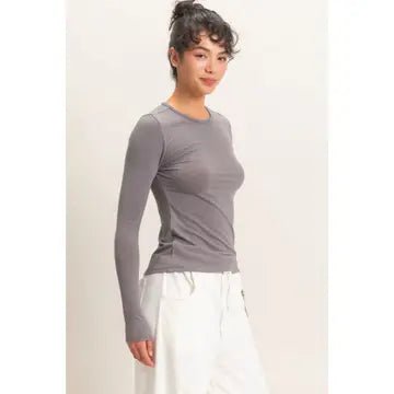A woman modeling a fitted, long-sleeve crew-neck athletic top in a medium gray color, paired with white drawstring pants, against a plain off-white studio background.
