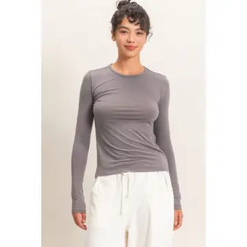 A woman modeling a fitted, long-sleeve crewneck top in a muted grayish-purple tone, paired with white drawstring pants, against a plain light beige studio background.
