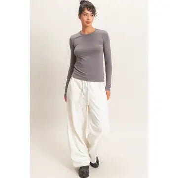 A woman modeling a tight-fitting long-sleeve crewneck top in gray, paired with loose white pants and black shoes, against a plain light background.