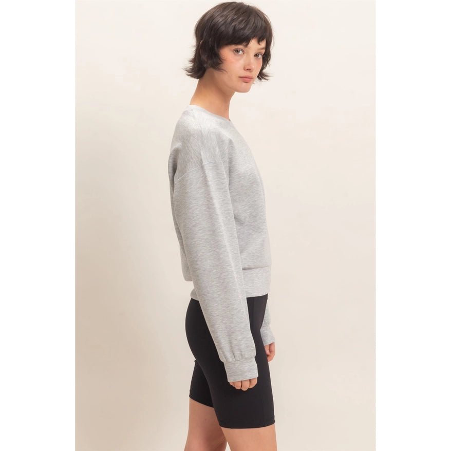 A woman shown in side profile wearing a light gray long-sleeve crewneck sweatshirt and black fitted shorts against a plain beige studio background.