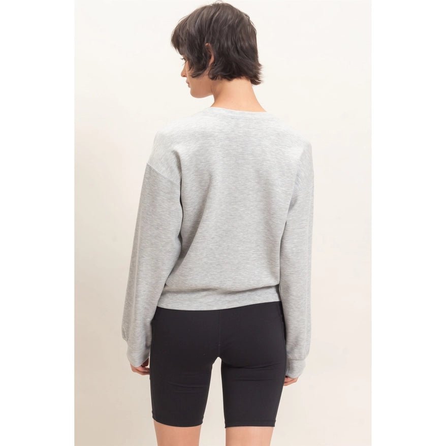 A woman shown from the back wearing a light gray heather crewneck sweatshirt and black bike shorts against a plain off-white studio background.