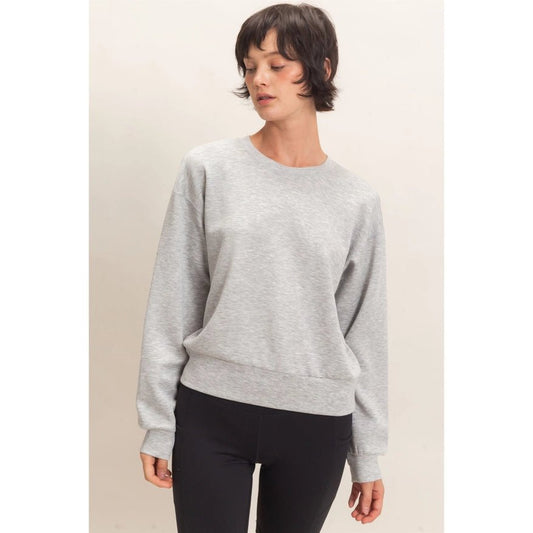 A woman wearing a light gray crewneck sweatshirt and black pants, standing against a plain beige studio background.