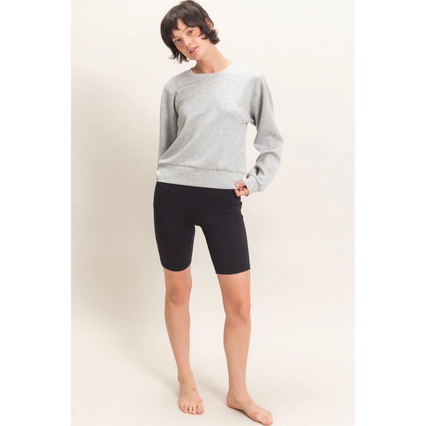 A woman modeling a light gray crewneck sweatshirt and black bike shorts against a plain off-white studio background.