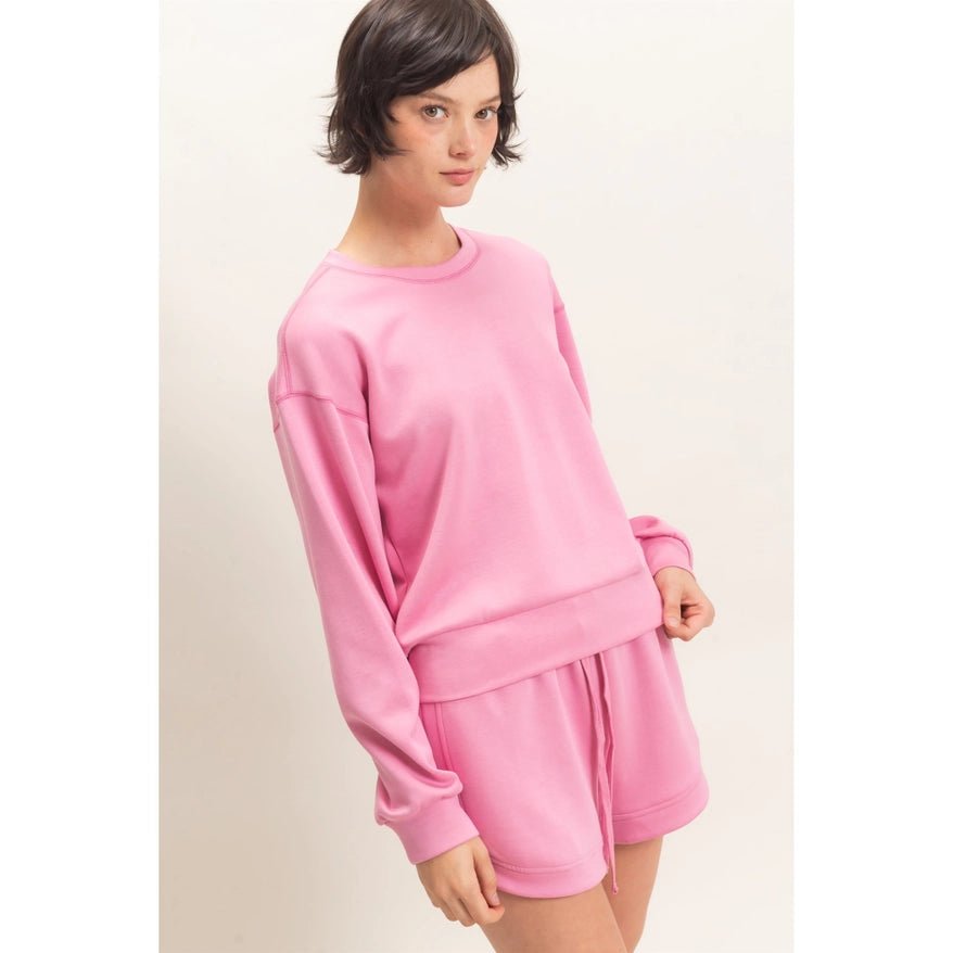 A woman modeling a matching pink lounge set consisting of a long-sleeve sweatshirt and drawstring shorts against a plain off-white background.