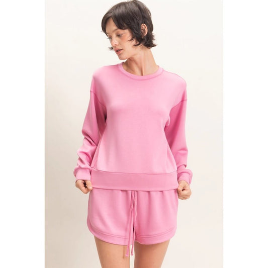 A woman modeling a matching pink lounge set consisting of a long-sleeve sweatshirt and drawstring shorts against a plain light background.