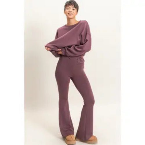 A model wearing a matching mauve-purple lounge set consisting of a long-sleeve top and flared pants, paired with tan slip-on shoes, against a plain light beige studio background.