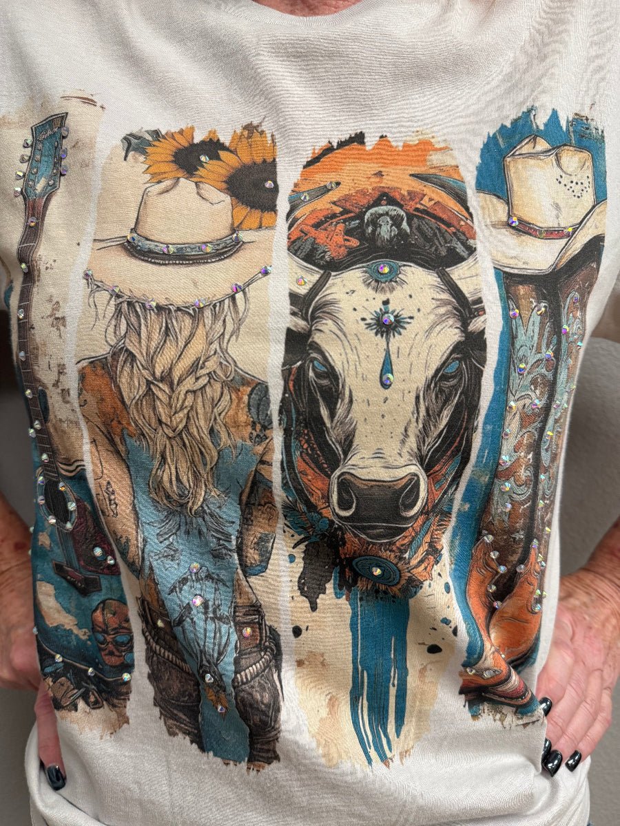 A close-up of a person wearing a light beige graphic T-shirt. The shirt features a large, detailed vertical western-themed print with a cow face, a woman in a cowboy hat, boots, and sunflowers in blue, orange, black, and beige tones.