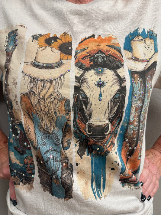 A close-up of a person wearing a light beige graphic T-shirt. The shirt features a large, detailed vertical western-themed print with a cow face, a woman in a cowboy hat, boots, and sunflowers in blue, orange, black, and beige tones.