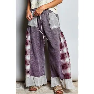 A person wearing loose, wide-leg patchwork pants with a purple and white checkered body, tie-dye side panels, and frayed white ruffle hems, paired with a cream top and sandals.
