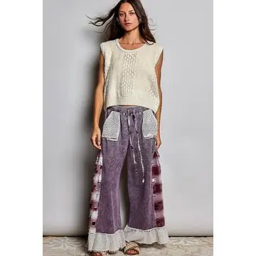 A woman modeling a sleeveless cream knit top and wide-leg purple pants with white lace trim and patterned side panels, standing against a plain light gray wall.