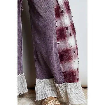 A close-up of a pair of wide-leg pants in a textured mauve-purple fabric, featuring a vertical patchwork panel with a distressed plaid pattern in dark red and white, finished with frayed white burlap-style cuffs.