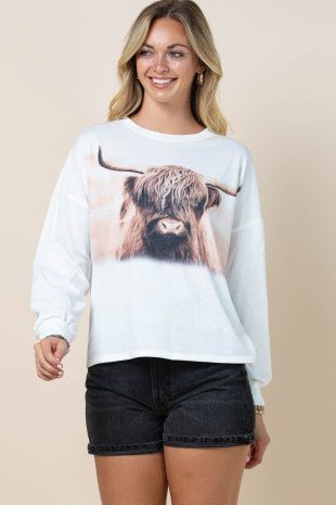 A woman modeling a white long-sleeve crewneck sweatshirt featuring a large graphic print of a Highland cow on the front, paired with black denim shorts against a solid beige background.