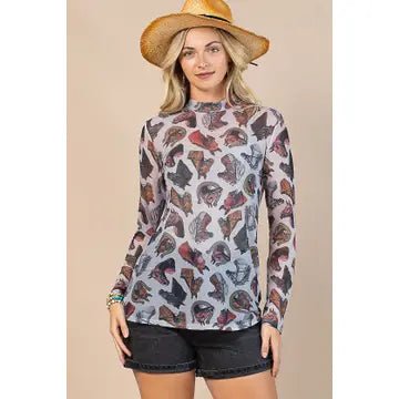 A woman modeling a long-sleeve mock neck top with an all-over butterfly print, paired with black denim shorts and a straw hat, set against a solid beige background.