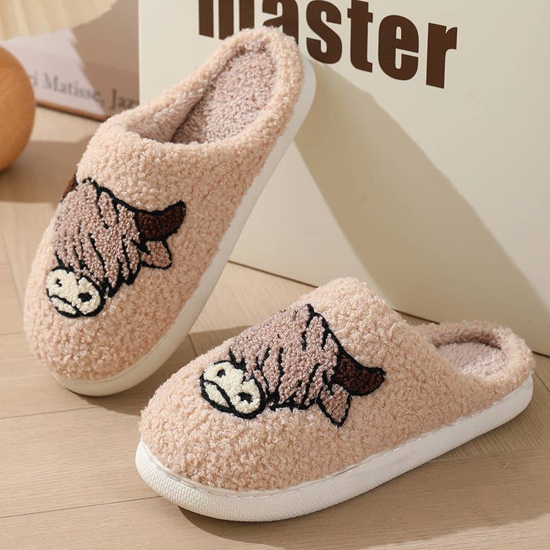 A pair of plush fleece slip-on house slippers in a light peach-pink color, featuring a cartoon cow embroidered on the top, resting on a light wood floor next to a box.