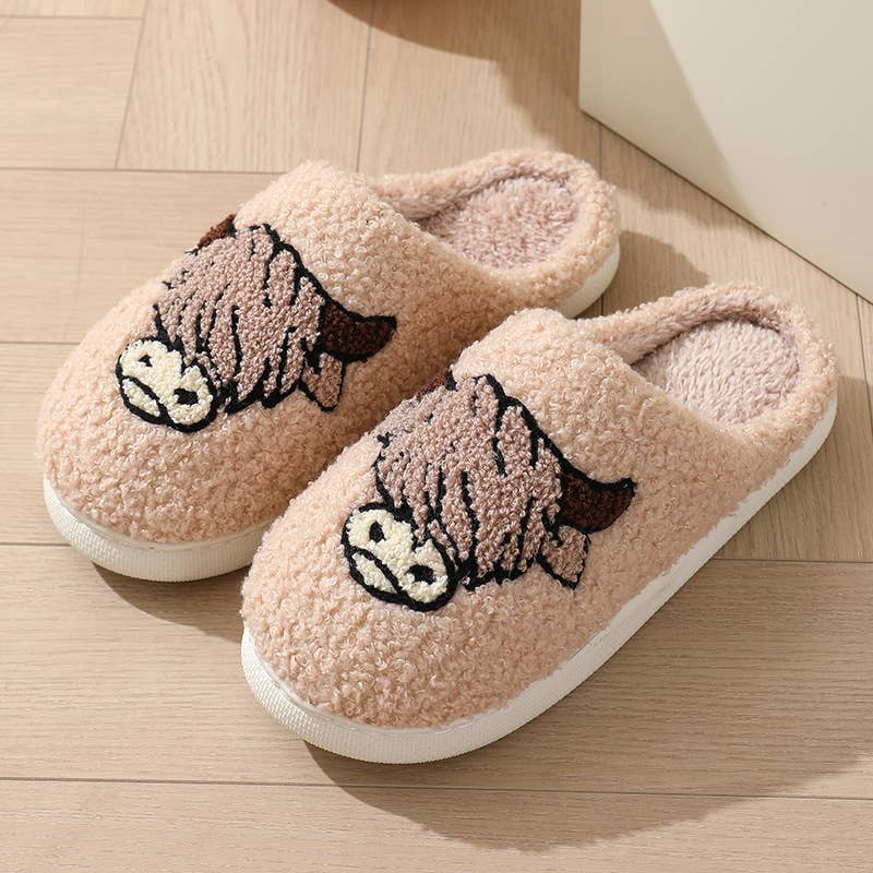 A pair of plush slip-on house slippers in a light peach-pink color, featuring embroidered cow faces on the top and white soles, shown on a light wood floor.