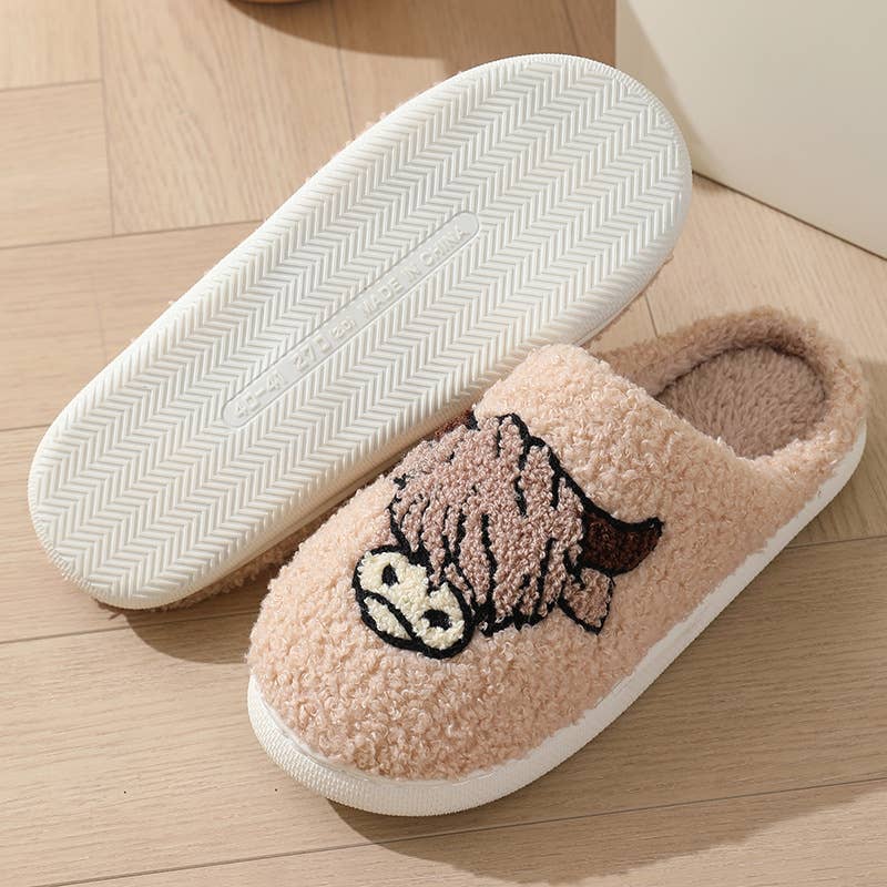 A pair of plush slip-on house slippers on a light wood floor. One slipper is shown from the top, featuring a textured beige sherpa body with a cartoon cow face embroidered on the side, and a white sole.