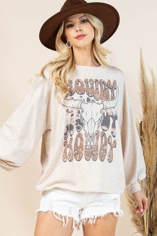 A woman wearing a beige oversized sweatshirt with a 'BOWDY BOWDY' cow skull graphic, styled with a brown wide-brim hat and white denim shorts against a neutral beige background.
