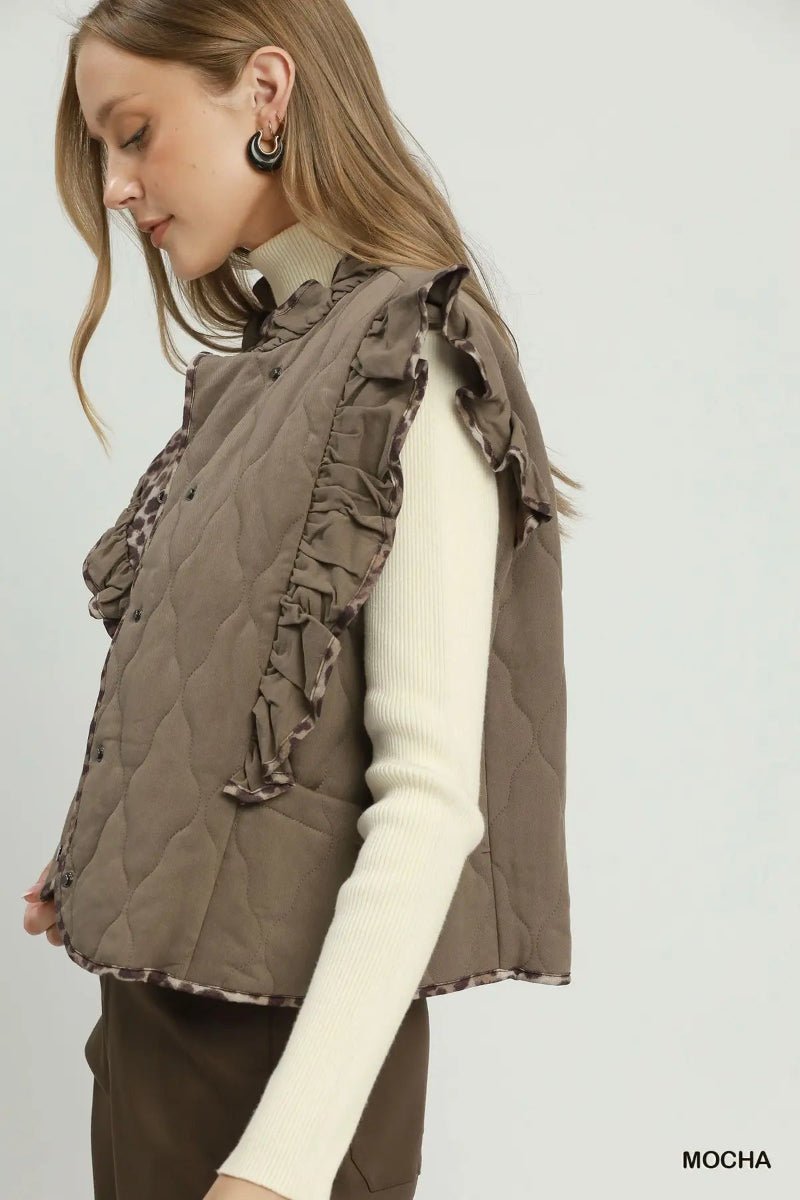 A woman wearing a sleeveless quilted vest in a warm brownish-tan color with ruffle trim, layered over a cream ribbed sweater. The image is shot against a plain light gray background with the word 'MOCHA' in the bottom right corner.