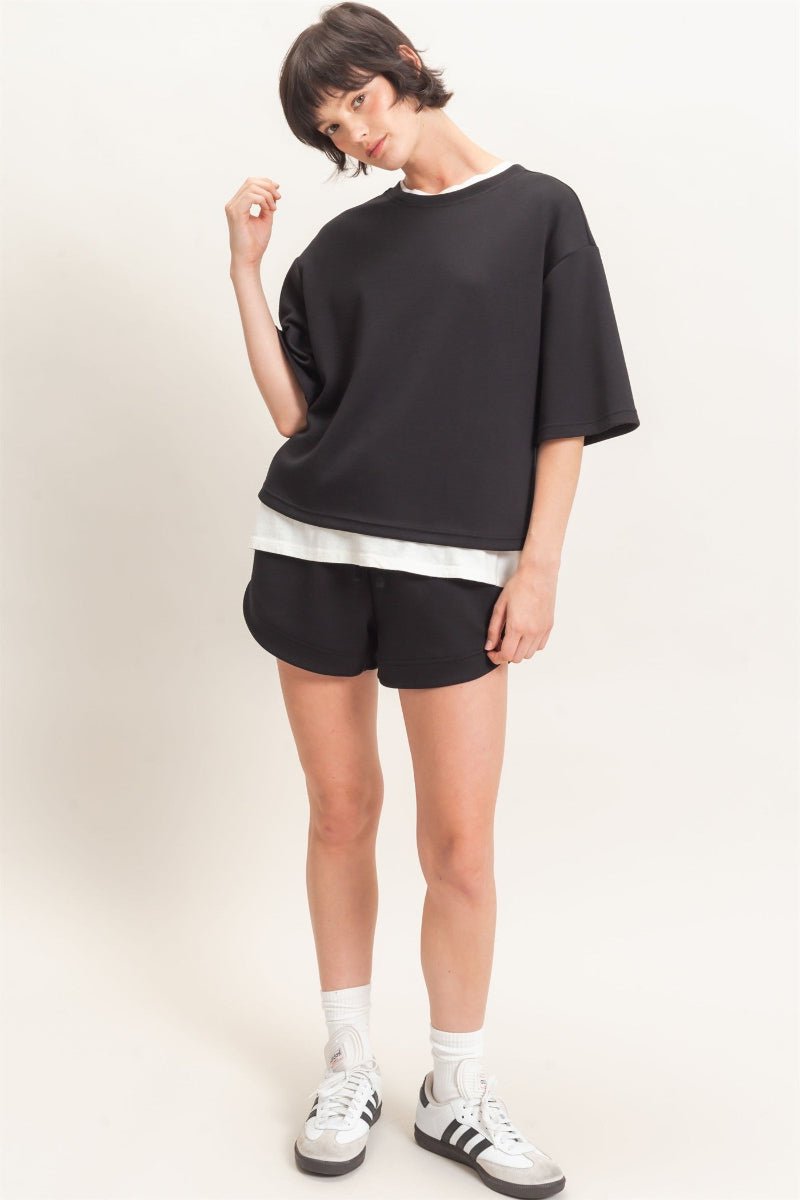 Person wearing a black oversized shirt and shorts with white sneakers on a plain background