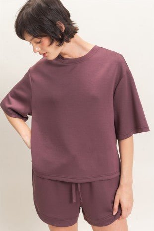 A woman wearing a matching mauve-brown lounge set consisting of a short-sleeve crewneck top and drawstring shorts, photographed against a plain light background.