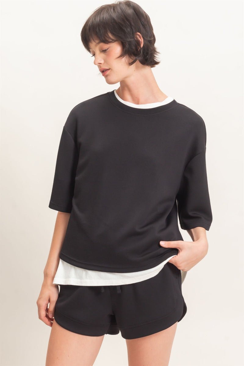 Person wearing a black sweater over a white shirt with black shorts on a plain background