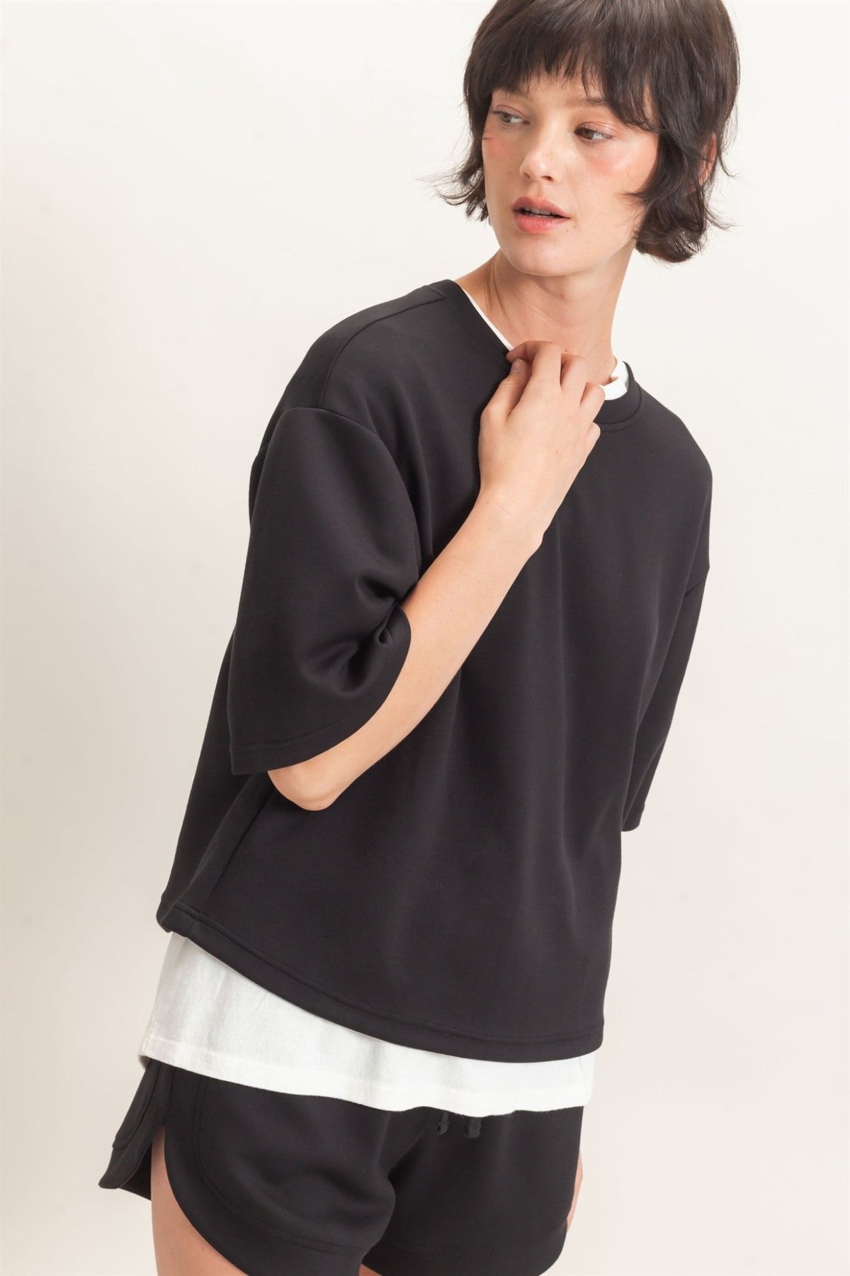 Person wearing a black oversized sweater over a white shirt with black shorts on a light background