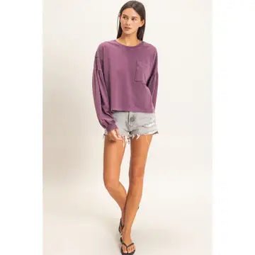 A woman modeling a long-sleeve, crewneck purple sweatshirt with a chest pocket, paired with light-wash denim shorts and black sandals, against a plain light gray studio background.