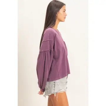A woman shown in side profile wearing a dusty purple cropped long-sleeve top with gathered sleeves and light-wash distressed denim shorts against a plain off-white background.