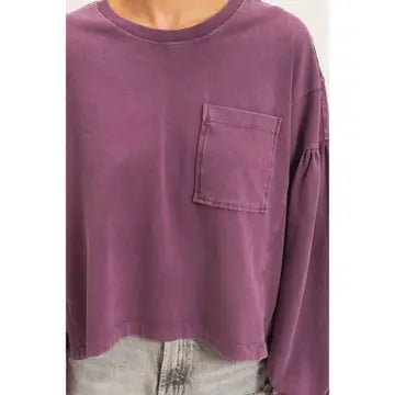 A close-up of a person wearing a cropped, long-sleeve t-shirt in a washed purple or mauve color, featuring a single chest pocket and a relaxed fit.