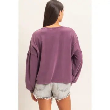 A woman shown from the back wearing a washed mauve-purple long-sleeve sweatshirt and light-wash denim shorts against a plain off-white background.