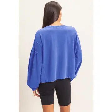 A woman viewed from the back wearing a cropped, oversized long-sleeve top in a medium royal blue color, paired with black bike shorts, against a plain off-white background.