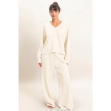 A woman modeling a cream-colored two-piece lounge set consisting of a long-sleeve V-neck top and matching wide-leg pants, set against a plain light beige studio background.