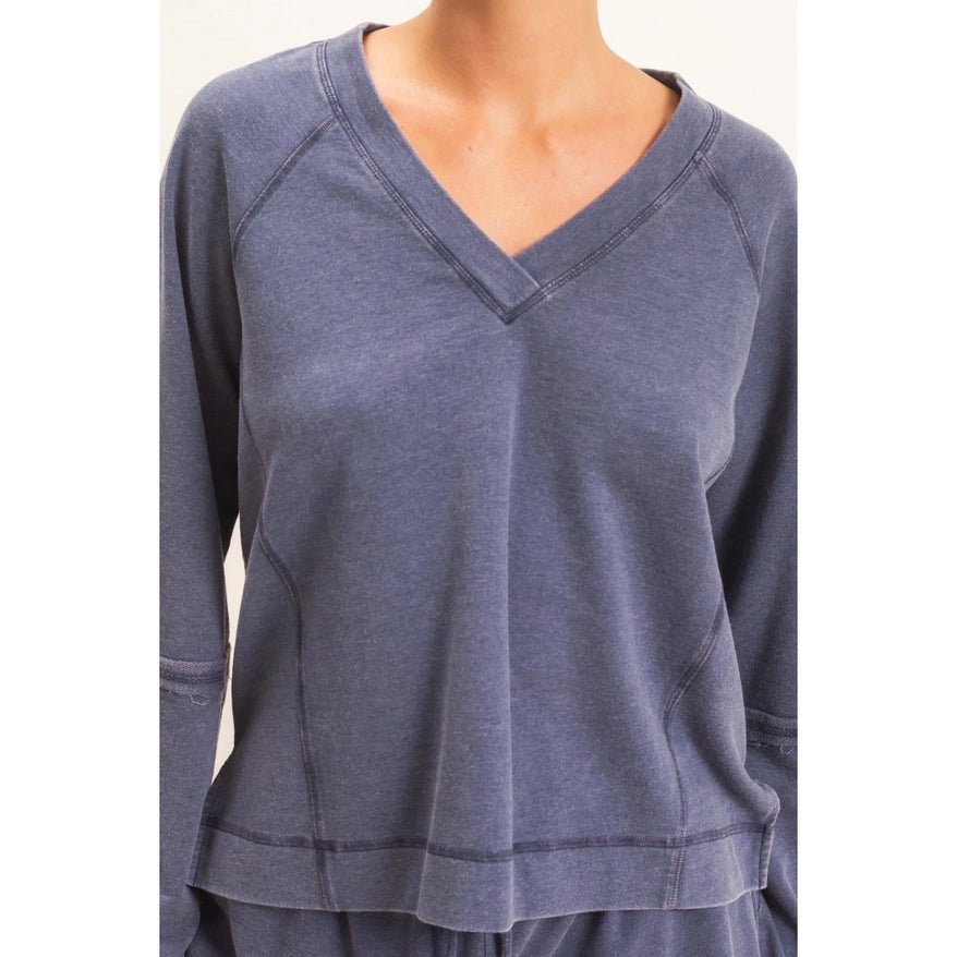 A close-up of a person wearing a slate blue-gray long-sleeve V-neck top, showing the chest, neckline, and raglan sleeves against a plain light background.