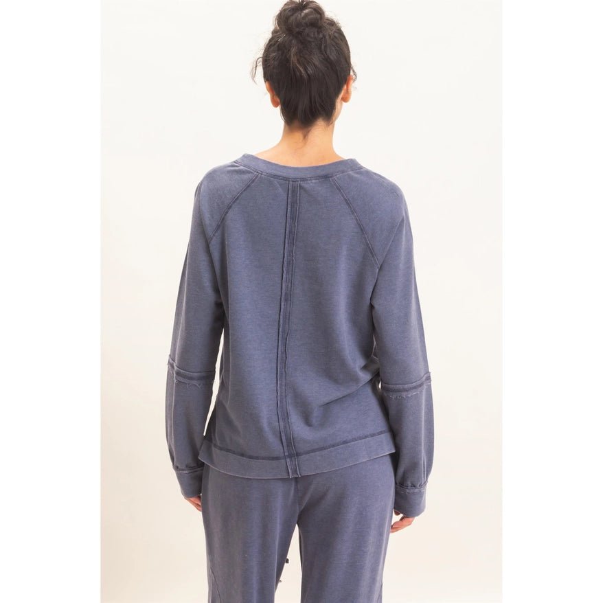 A woman viewed from behind wearing a heathered slate blue long-sleeve sweatshirt and matching sweatpants against a plain off-white studio background.