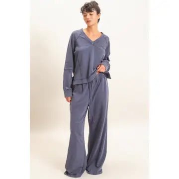 A woman modeling a matching two-piece loungewear set in a dusty slate blue-gray color, featuring a long-sleeve V-neck top and wide-leg pants, against a plain light beige studio background.