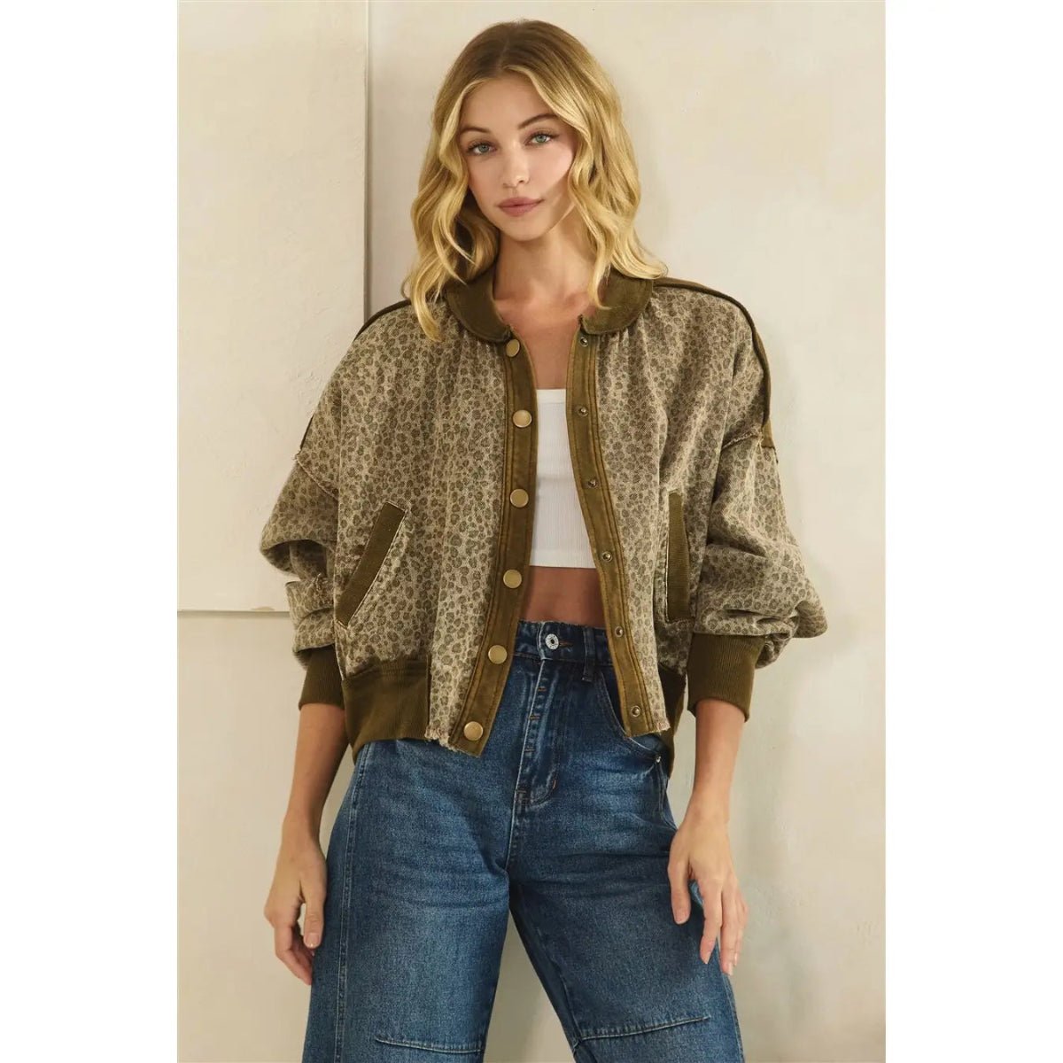A woman modeling a cropped leopard-print bomber jacket with olive green ribbed trim and gold-tone buttons, paired with a white top and blue jeans against a light beige wall.