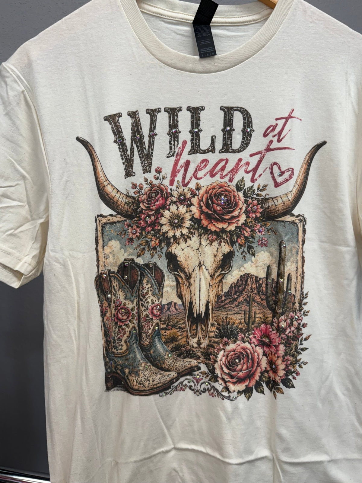 An off-white short-sleeve graphic t-shirt hanging on a wooden hanger against a dark gray background. The graphic features a western theme with a skull, floral elements, cowboy boots, and the text 'WILD at heart'.