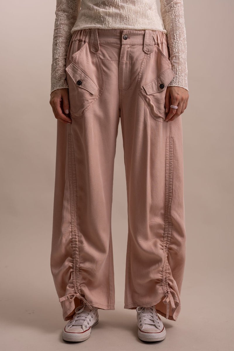 A person wearing wide-leg dusty pink cargo pants with front flap pockets and gathered ruffle details at the hems, paired with a cream textured long-sleeve top and white sneakers, against a plain beige studio background.