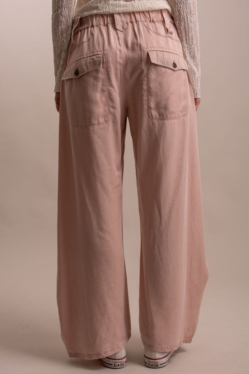 A person shown from the back wearing dusty pink wide-leg pants with an elastic waistband and flap pockets, paired with a cream knit top and white sneakers against a plain beige studio background.
