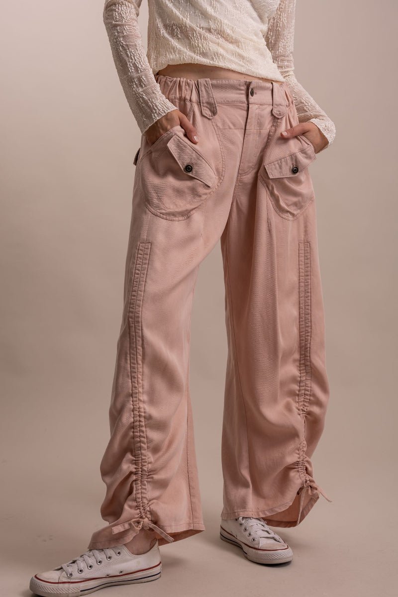 A person wearing dusty pink wide-leg cargo pants with side ruching, pleats, and drawstring details, styled with a cream lace-up top and white sneakers against a solid beige studio background.