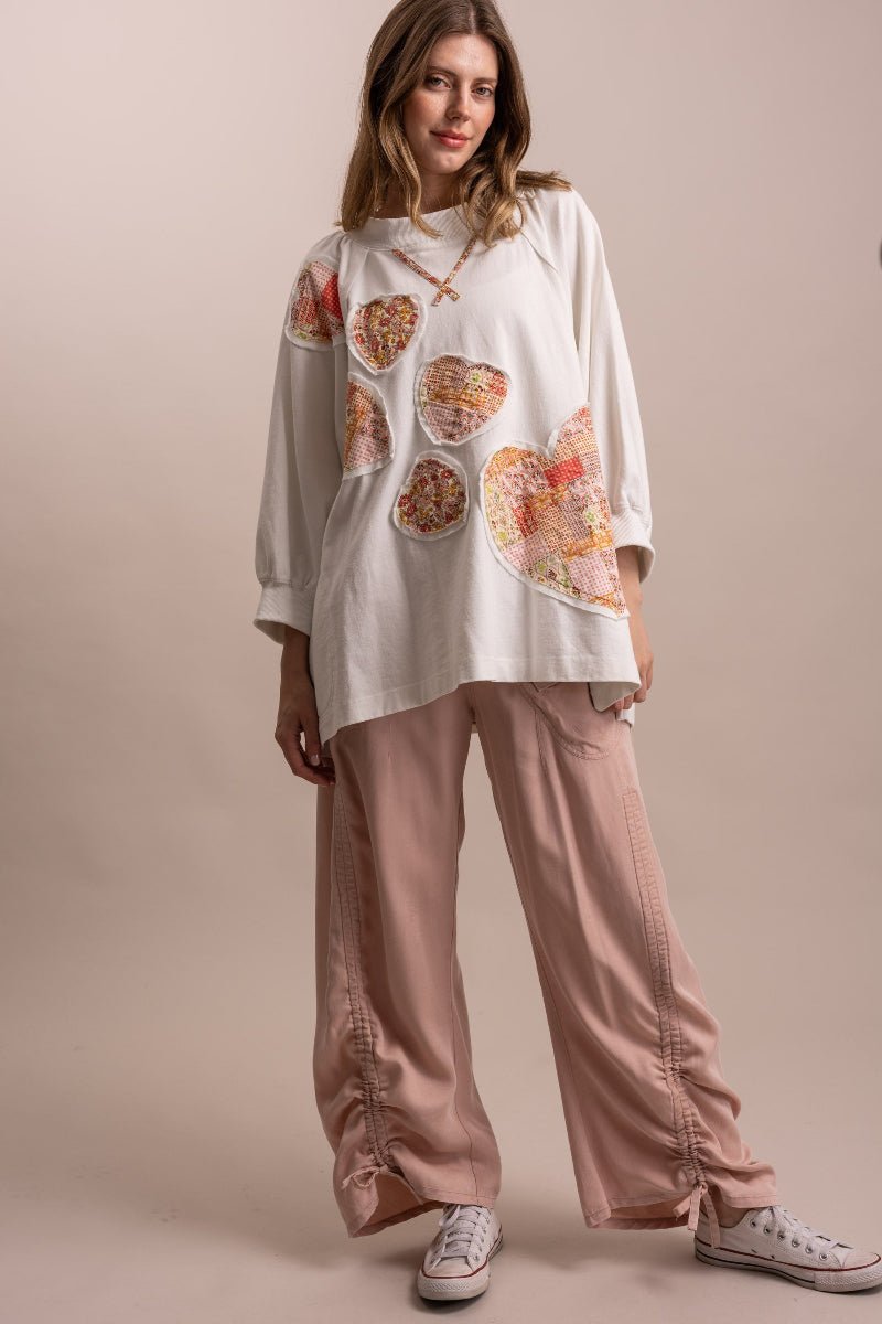 A woman modeling a white long-sleeve top with colorful heart-shaped patchwork prints, paired with dusty pink wide-leg pants and white sneakers, against a solid beige studio background.