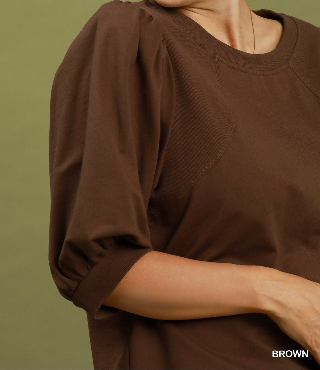 A close-up of a person wearing a brown short-sleeve top with voluminous elbow-length puff sleeves and a crew neckline, against a muted olive-green background. The word 'BROWN' appears in the bottom right corner.
