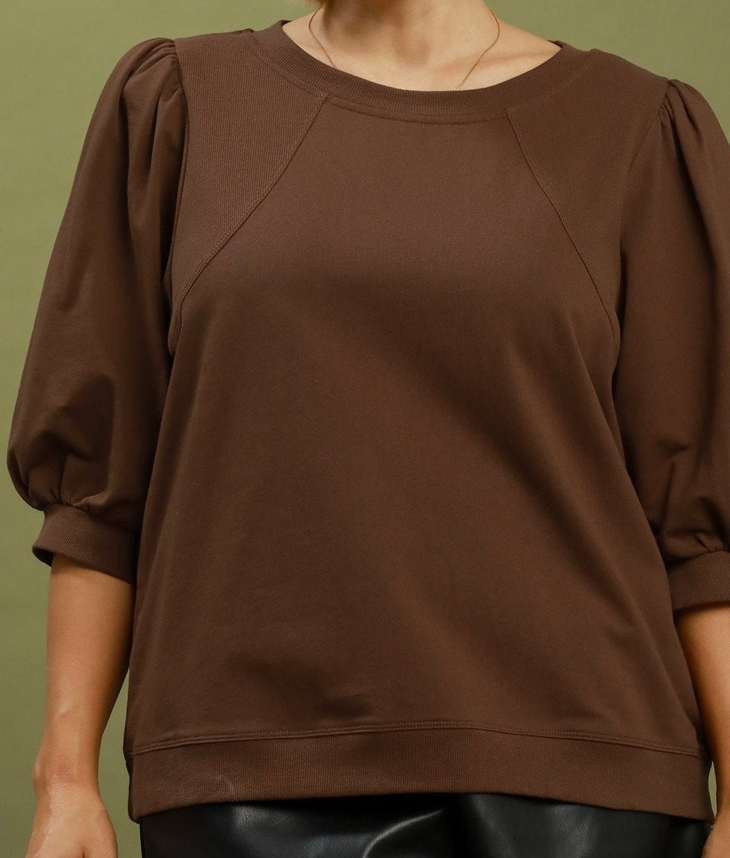 A person wearing a brown crewneck top with voluminous elbow-length puff sleeves and a relaxed silhouette, shown against a muted olive background.