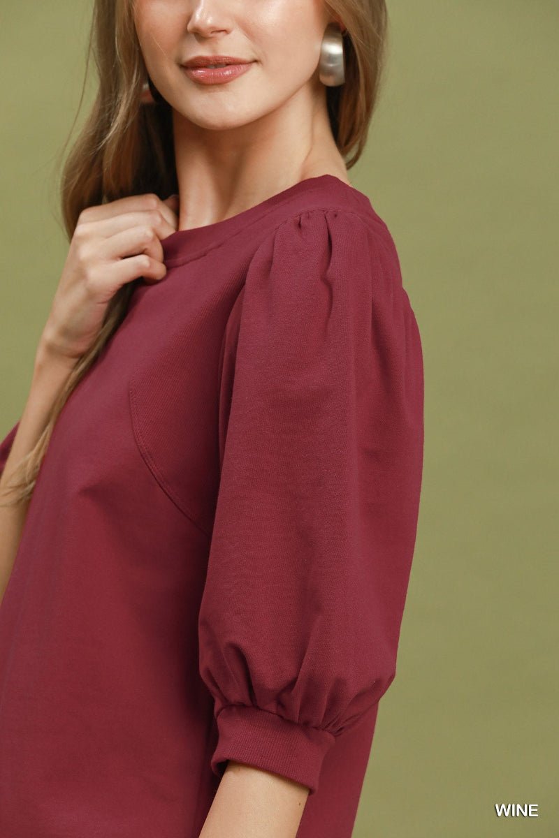 A close-up of a woman wearing a wine-colored puff-sleeve blouse against an olive green background.