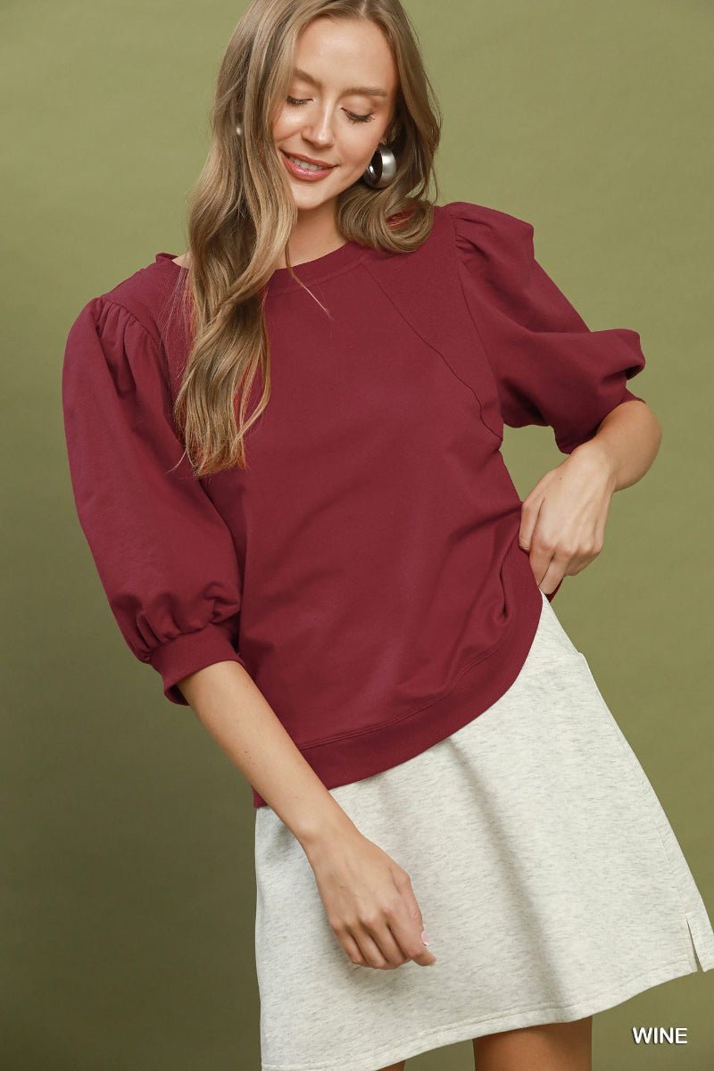 A woman wearing a wine-colored puff-sleeve top and a light gray skirt, posing against a solid olive green background. The word 'WINE' is printed in white in the bottom right corner.