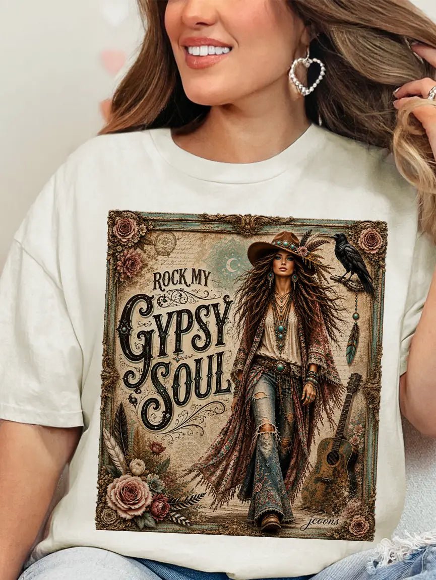A woman wearing a cream-colored graphic t-shirt with a detailed western-themed print that reads 'ROCK MY GYPSY SOUL'. The print features an illustration of a woman in a cowboy hat, a raven, a guitar, and roses.