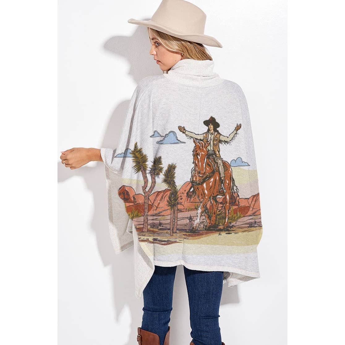 A woman wearing a light gray poncho-style wrap with a large western desert scene print, paired with blue jeans, brown boots, and a beige hat, shown from the back against a white studio background.