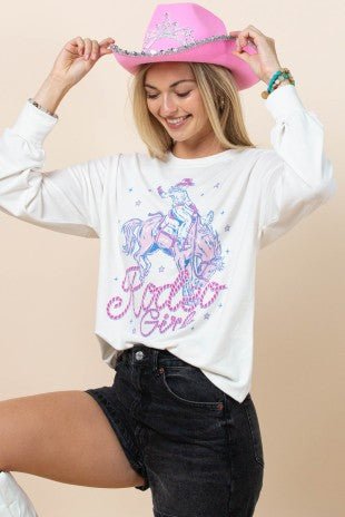 A woman wearing a white long-sleeve graphic t-shirt with a pink and blue 'Ride Like a Girl' cowboy graphic, paired with black denim shorts and a pink cowboy hat, posing against a solid beige background.